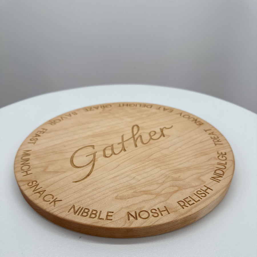 A round cherry hardwood serving board engraved with the word 'Gather' in the center and words like 'snack', 'nibble', 'nosh', 'relish', and 'indulge' around the edge, shown on a white round surface against a light gray background.