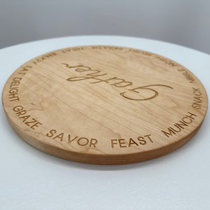 A round cherry hardwood serving board engraved with words like 'Gather', 'Feast', 'Munch', and 'Savor'. 