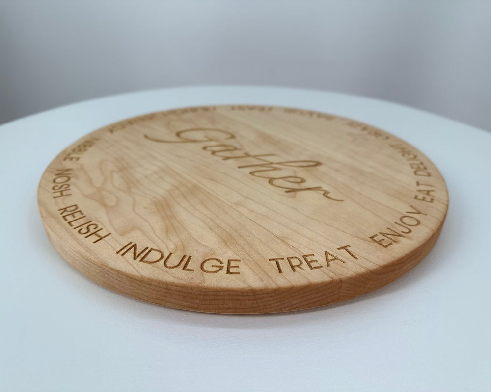 A round cherry hardwood serving board engraved with the word 'Gather' in the center and the words 'ENJOY', 'TREAT', 'INDULGE', 'RELISH', and 'GATHER' around the edge, shown on a white surface.