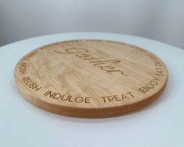 A round cherry hardwood serving board engraved with the word 'Gather' in the center and the words 'ENJOY', 'TREAT', 'INDULGE', 'RELISH', and 'GATHER' around the edge, shown on a white surface.