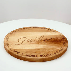 A round cherry hardwood serving board engraved with the word 'Gather' in the center and various spirits listed around the edge.