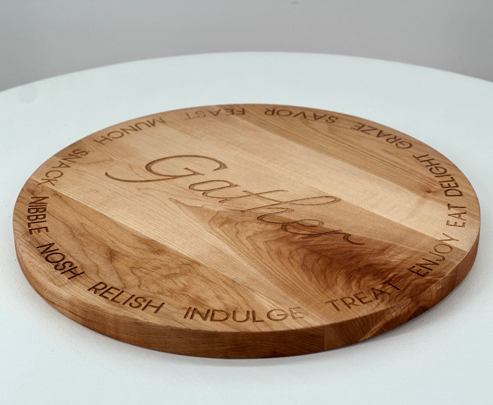 A round cherry wood serving board engraved with the word 'Gather' in the center and various positive words around the edge.