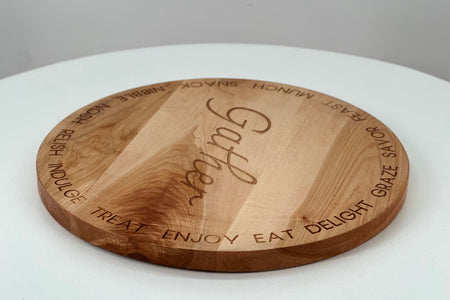 A round cherry hardwood serving board engraved with the word 'Gather' in the center and words like 'Treat', 'Enjoy', 'Eat', 'Delight', and 'Graze' around the edge.
