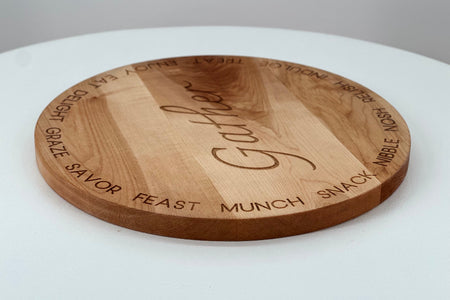 A round wooden serving board engraved with the word 'Gather' in the center and words like 'snack', 'munch', 'feast', and 'gather' around the edge, shown on a white surface.