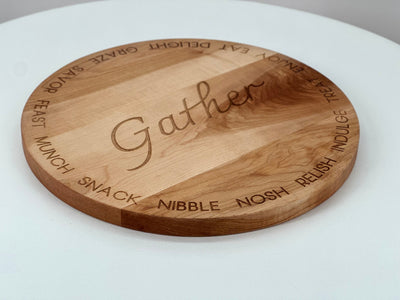 A round cherry hardwood serving board engraved with the word 'Gather' in the center and surrounding words like munch, snack, nibble, and relish.