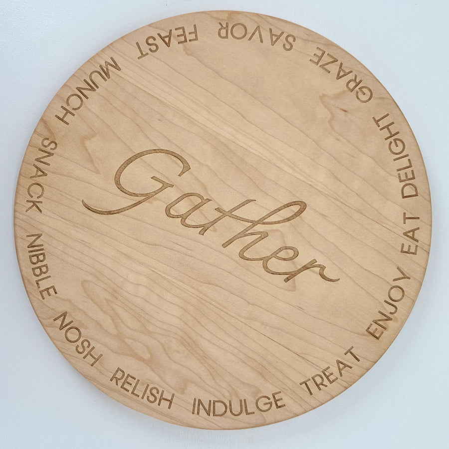 Overhead view of round cherry hardwood serving tray with engraved words like feast munch snack nibble nosh relish indulge treat enjoy eat delight graze on the boarder of the piece and the word Gather in the Center.