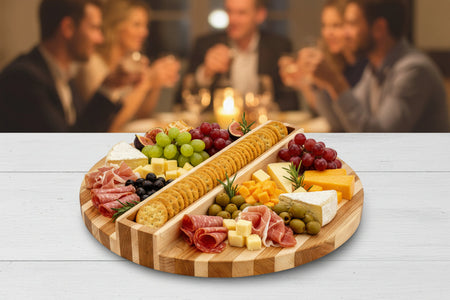 Wooden charcuterie board with meats, cheeses, fruits and a special compartment for crackers (known as the Cracker Keeper)on a table with people in the background.